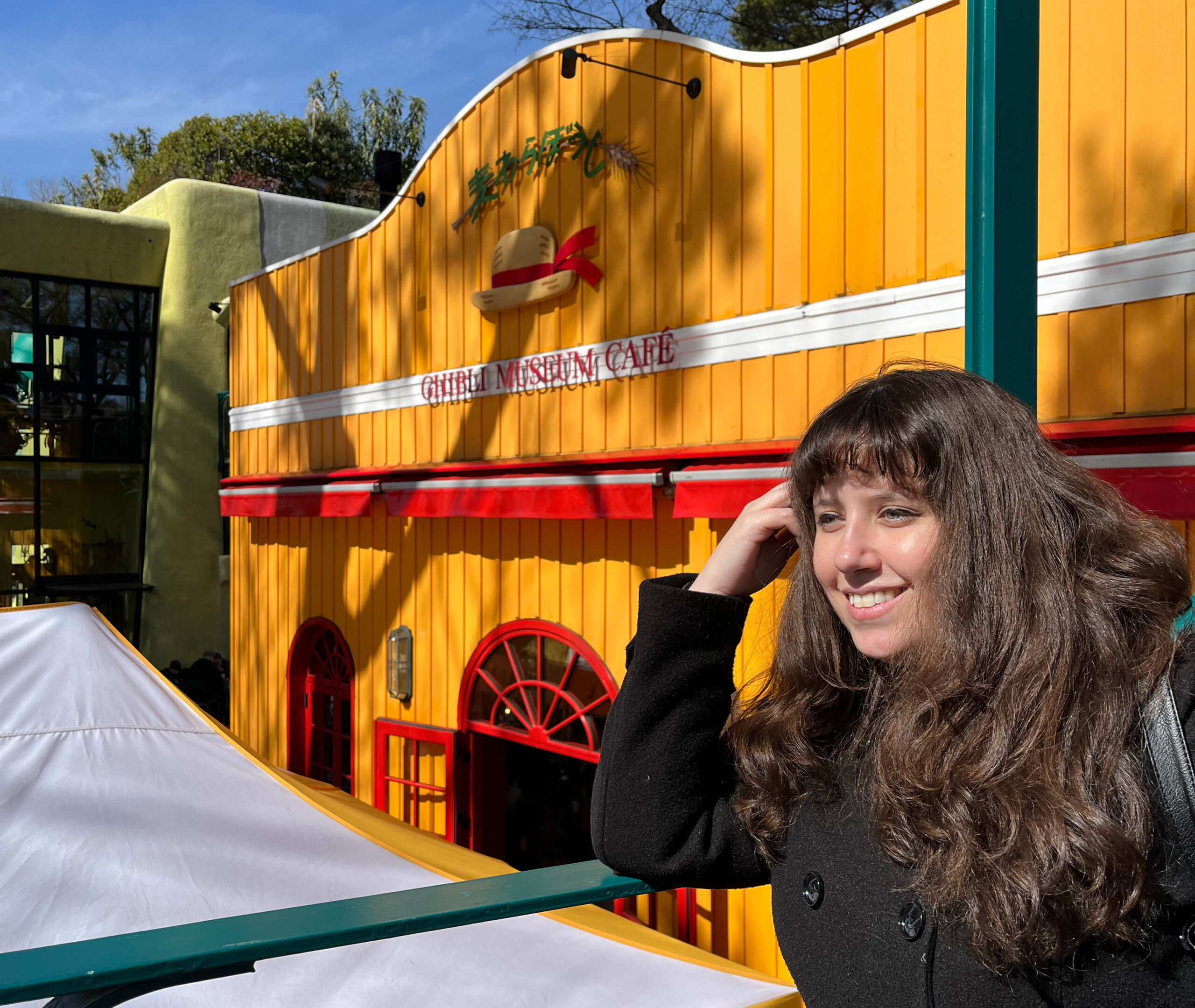 A woman in a black coat stands smiling outside the Ghibli Museum Cafe in Japan. The building is painted bright yellow with a green roof, and trees are visible in the background.